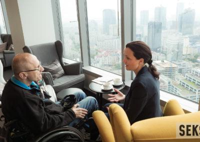 Uczestnicy konferencji w trakcie rozmowy podczas przerwy kawowej. Kobieta siedząca na fotelu oraz mężczyzna na wózku.