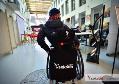 Zdjęcie z robione od tyłu. Młody mężczyzna na wózku inwalidzkim, uczestnik konferencji. Na rączkach wózka wisi torba z logiem projektu Sekson.