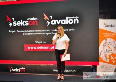 Agata Niemiec na scenie, mówi do mikrofonu, za nią duży ekran z logo projektu Sekson i Fundacji Avalon.