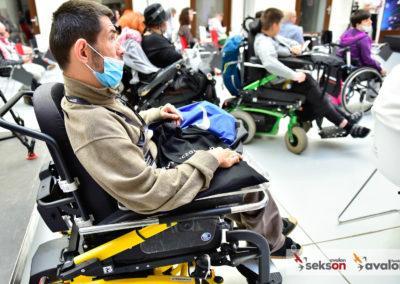 Zdjęcie zrobione z boku, na pierwszym planie jeden z uczestników konferencji - mężczyzna na wózku inwalidzkim, w maseczce. W tle pozostali uczestnicy.